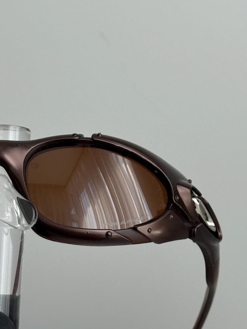 May include: Close-up of a pair of brown-tinted sunglasses with a dark brown frame. The sunglasses have a curved design and a silver accent on the temple. The lenses are polarized.