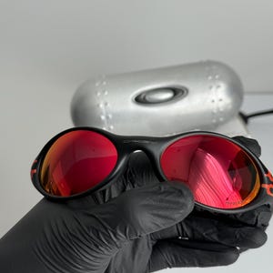May include: Black sunglasses with red lenses. The sunglasses have a black frame with orange accents. The lenses are reflective. The sunglasses are held by a gloved hand. A silver case is in the background.