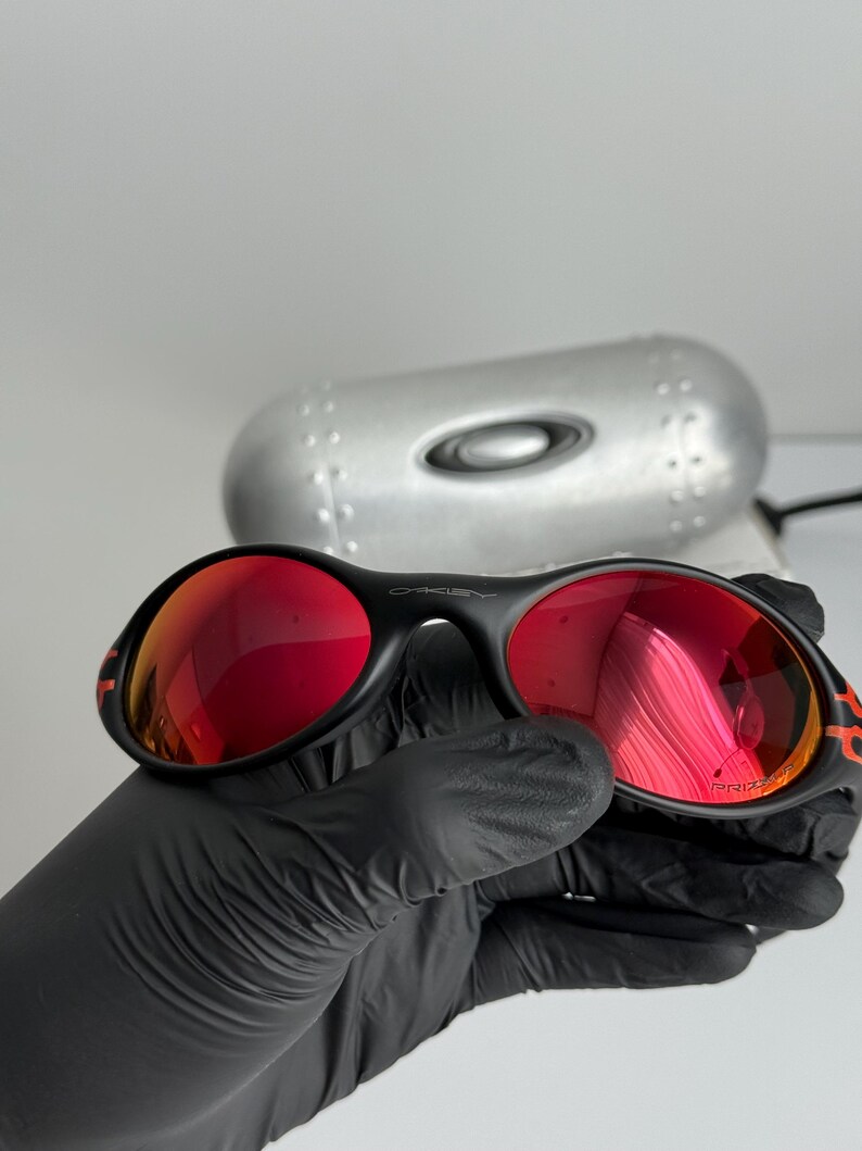May include: Black sunglasses with red lenses. The sunglasses have a black frame with orange accents. The lenses are reflective. The sunglasses are held by a gloved hand. A silver case is in the background.