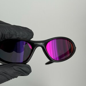 May include: Black Oakley sunglasses with a curved frame and pink and purple lenses. The Oakley logo is visible on the top of the frame. The sunglasses are held by a gloved hand.