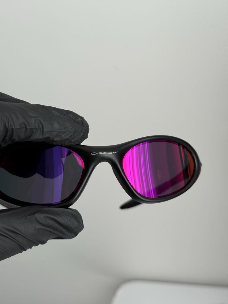 May include: Black Oakley sunglasses with a curved frame and pink and purple lenses. The Oakley logo is visible on the top of the frame. The sunglasses are held by a gloved hand.