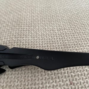 May include: Close-up of black sunglasses with the text "MADE IN U.S.A." printed on the arm. The sunglasses are set against a neutral, textured background.