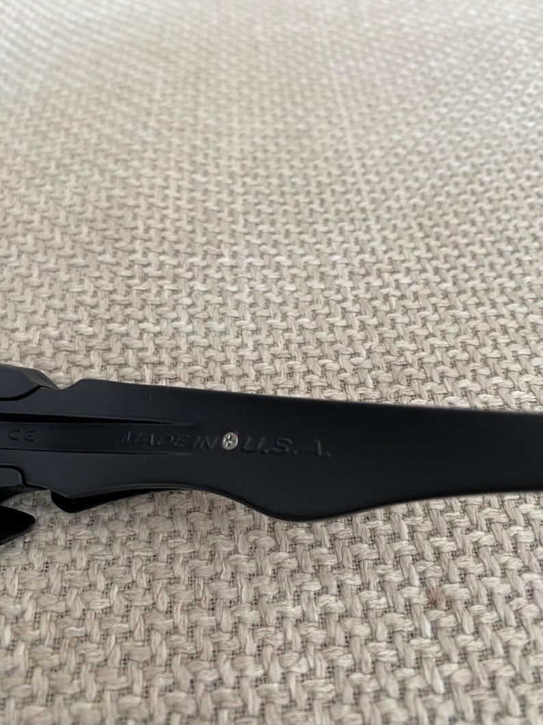 May include: Close-up of black sunglasses with the text "MADE IN U.S.A." printed on the arm. The sunglasses are set against a neutral, textured background.
