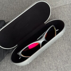 May include: Silver-framed sunglasses with red-tinted lenses rest inside an open, silver case with a black interior. The case is oval-shaped and has a textured surface. The sunglasses have a sleek, modern design.