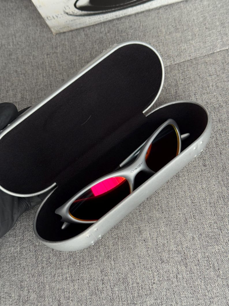 May include: Silver-framed sunglasses with red-tinted lenses rest inside an open, silver case with a black interior. The case is oval-shaped and has a textured surface. The sunglasses have a sleek, modern design.