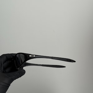 K&ouml;nnte beinhalten: Schwarze Sonnenbrille mit einem schlanken, modernen Design. Die Sonnenbrille hat einen gebogenen Rahmen und dunkle Gl&auml;ser. Die B&uuml;gel sind d&uuml;nn und reichen bis zum Ohr. Die Sonnenbrille wird von einer behandschuhten Hand gehalten.