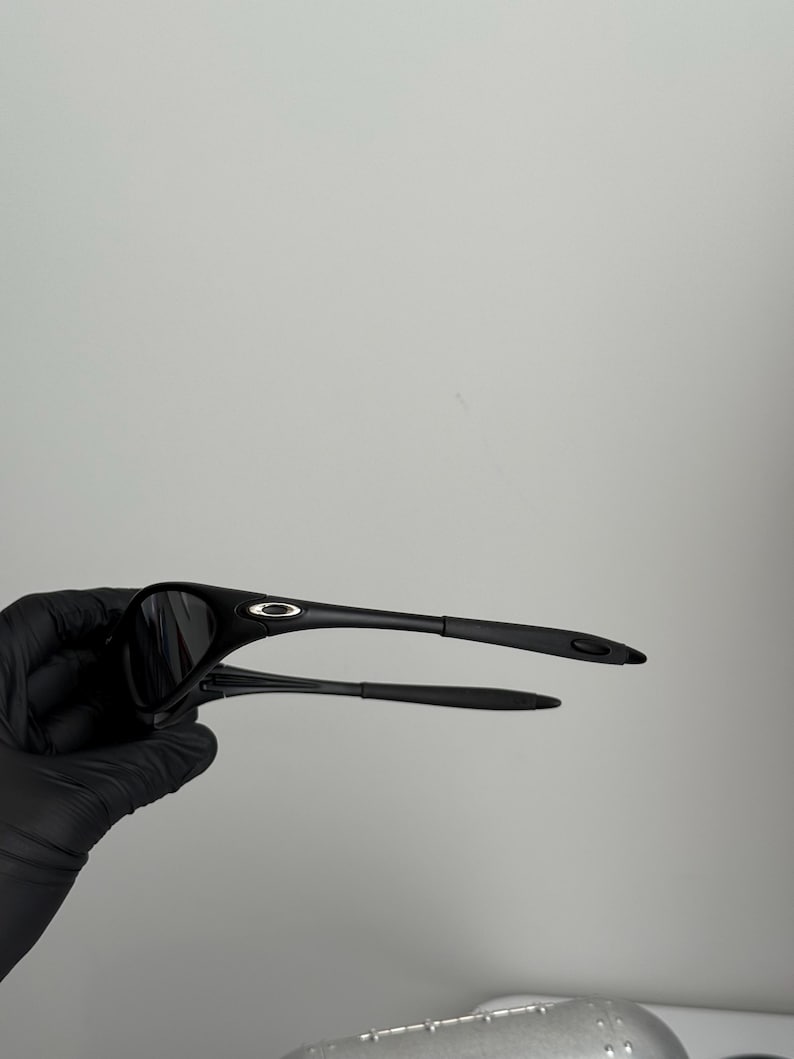 K&ouml;nnte beinhalten: Schwarze Sonnenbrille mit einem schlanken, modernen Design. Die Sonnenbrille hat einen gebogenen Rahmen und dunkle Gl&auml;ser. Die B&uuml;gel sind d&uuml;nn und reichen bis zum Ohr. Die Sonnenbrille wird von einer behandschuhten Hand gehalten.