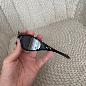 Puede incluir: Gafas de sol negras con montura curvada y lentes oscuras. Las gafas de sol tienen un peque&ntilde;o logotipo dorado en la patilla. Se sostienen en una mano.