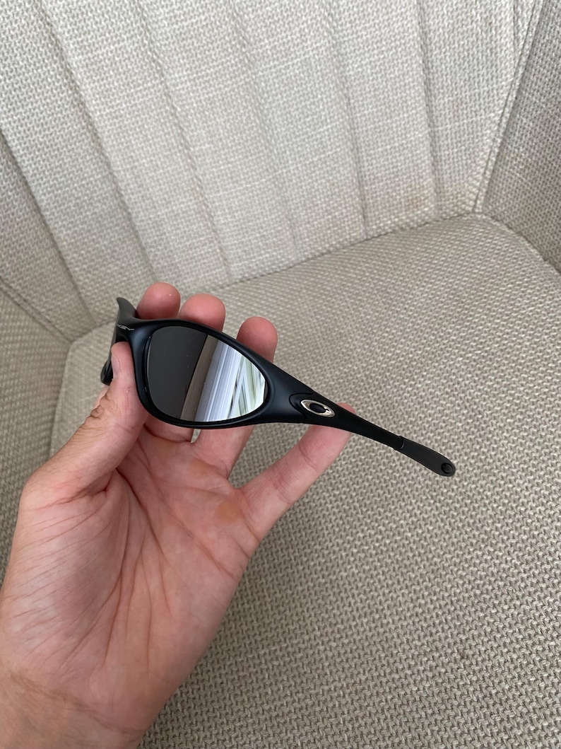 Puede incluir: Gafas de sol negras con montura curvada y lentes oscuras. Las gafas de sol tienen un peque&ntilde;o logotipo dorado en la patilla. Se sostienen en una mano.