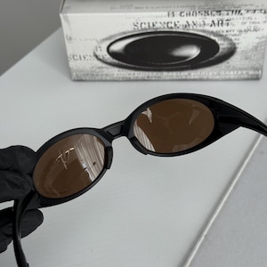 Puede incluir: Gafas de sol ovaladas negras con lentes marrones. Las gafas de sol tienen un dise&ntilde;o elegante y moderno. La imagen tambi&eacute;n muestra una caja blanca con el texto "SCIENCE AND ART" y un estuche plateado.