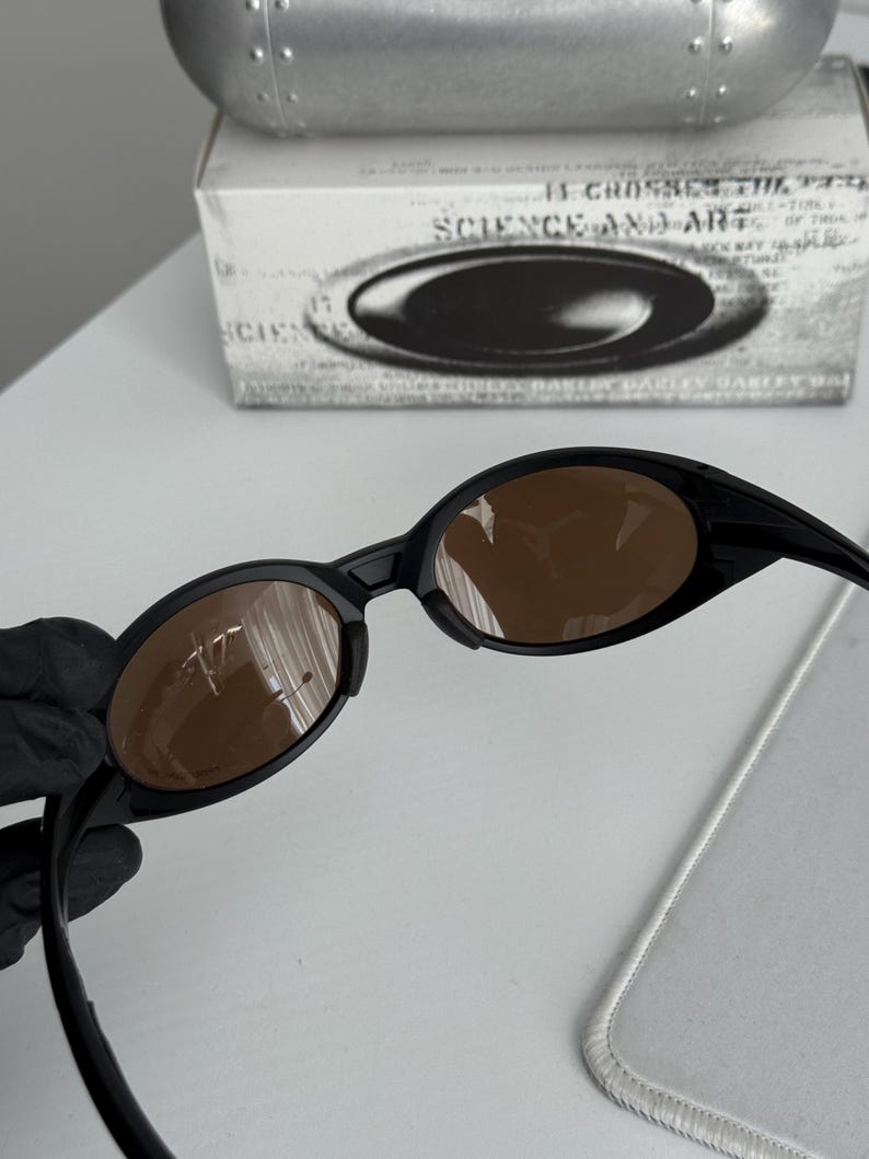 Puede incluir: Gafas de sol ovaladas negras con lentes marrones. Las gafas de sol tienen un dise&ntilde;o elegante y moderno. La imagen tambi&eacute;n muestra una caja blanca con el texto "SCIENCE AND ART" y un estuche plateado.