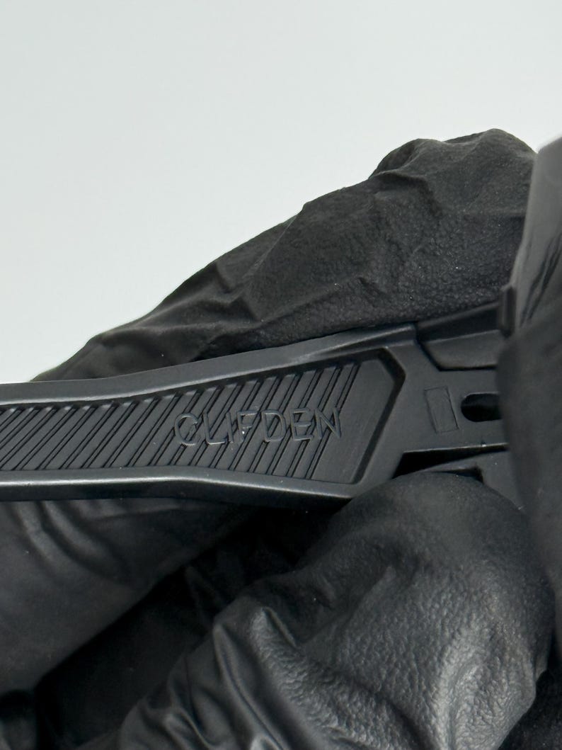 K&ouml;nnte beinhalten: Nahaufnahme eines schwarzen Teppichmessers mit dem Wort "CLIFDEN" auf dem Griff. Das Messer wird von einer behandschuhten Hand vor wei&szlig;em Hintergrund gehalten. Der Griff hat ein geriffeltes Design f&uuml;r besseren Halt.