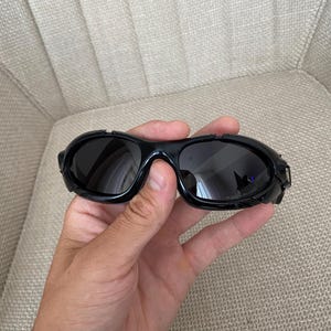 May include: Black wraparound sunglasses with dark lenses and thick, glossy frames. The sunglasses are held in a hand. The background is neutral.