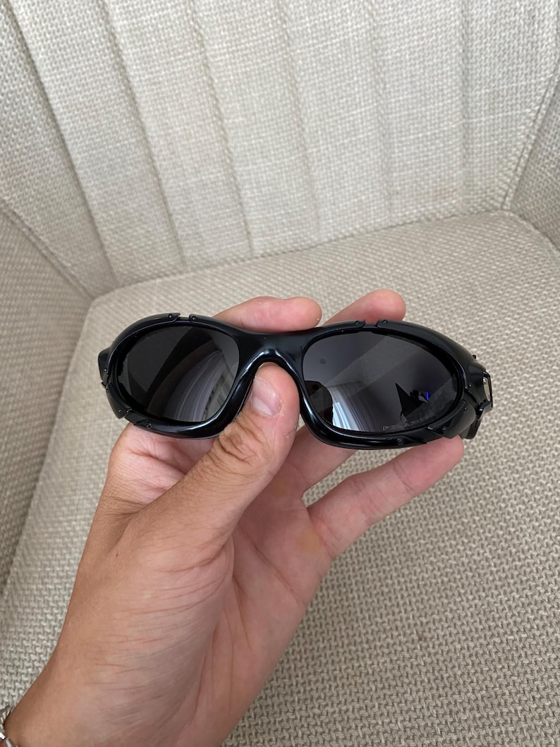 May include: Black wraparound sunglasses with dark lenses and thick, glossy frames. The sunglasses are held in a hand. The background is neutral.