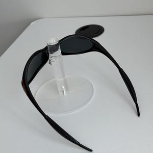 May include: Black sunglasses with a sleek, curved design. The sunglasses have dark lenses and a thin frame with a red accent on the temple. The sunglasses are displayed on a clear stand.
