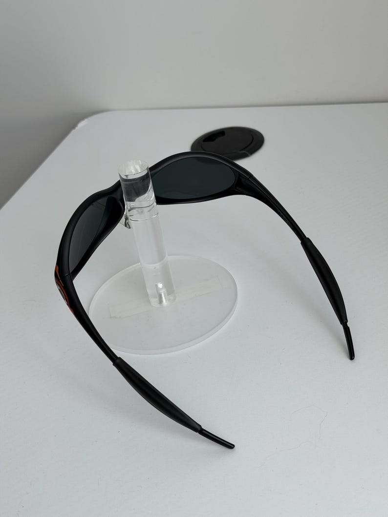 May include: Black sunglasses with a sleek, curved design. The sunglasses have dark lenses and a thin frame with a red accent on the temple. The sunglasses are displayed on a clear stand.