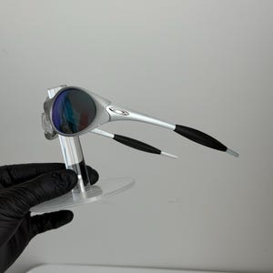 May include: Silver sunglasses with dark lenses. The sunglasses have a sleek design with black accents on the arms. The lenses reflect blue and purple. The sunglasses are displayed on a clear stand.
