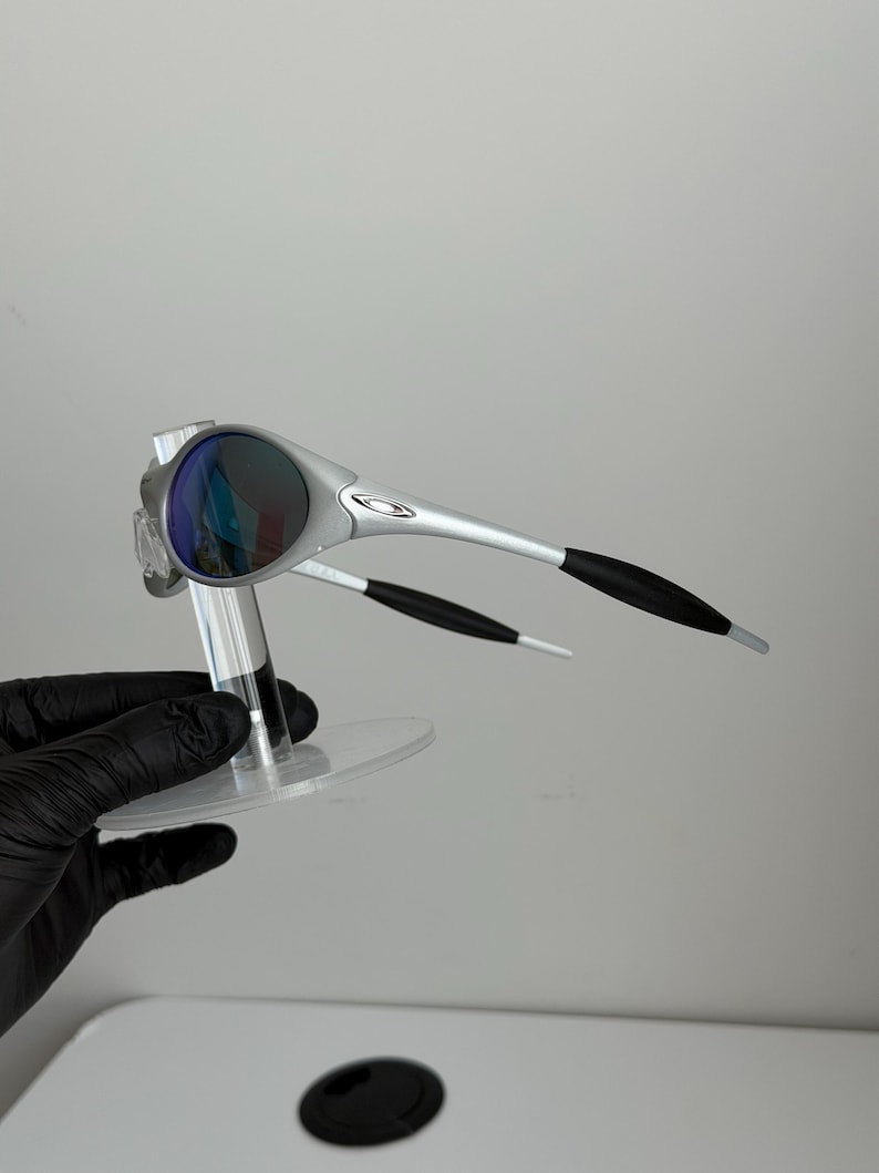 May include: Silver sunglasses with dark lenses. The sunglasses have a sleek design with black accents on the arms. The lenses reflect blue and purple. The sunglasses are displayed on a clear stand.