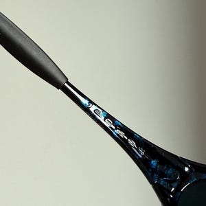 Puede incluir: Primer plano de unas gafas de sol. La montura negra tiene un patr&oacute;n azul y negro. La patilla de las gafas de sol tiene una empu&ntilde;adura de goma negra. Las lentes son oscuras.