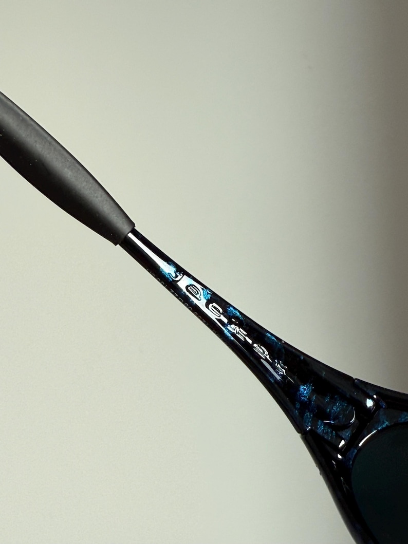 Puede incluir: Primer plano de unas gafas de sol. La montura negra tiene un patr&oacute;n azul y negro. La patilla de las gafas de sol tiene una empu&ntilde;adura de goma negra. Las lentes son oscuras.