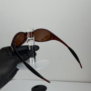 May include: Brown oval sunglasses with a dark red frame and brown lenses. The temples are black with red tips. The sunglasses are displayed on a clear stand.