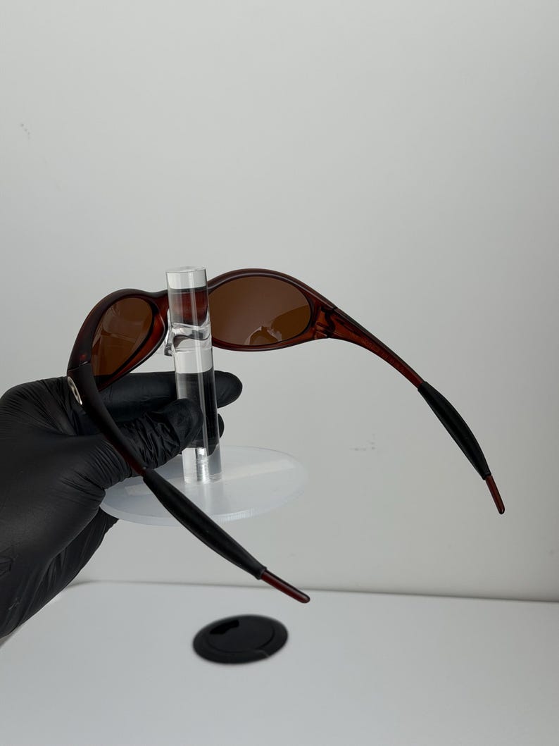 May include: Brown oval sunglasses with a dark red frame and brown lenses. The temples are black with red tips. The sunglasses are displayed on a clear stand.