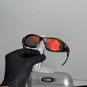 May include: Black framed sunglasses with red mirrored lenses. The sunglasses have a sporty design with a curved frame and a red accent on the temple. The lenses reflect a bright orange color.