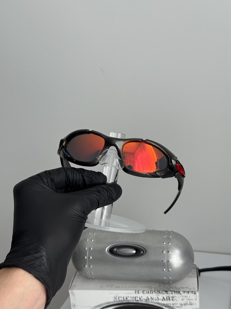 May include: Black framed sunglasses with red mirrored lenses. The sunglasses have a sporty design with a curved frame and a red accent on the temple. The lenses reflect a bright orange color.