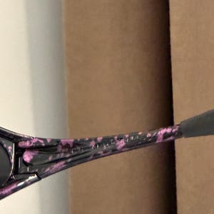 May include: Black sunglasses with a speckled pink and black pattern on the arms. The sunglasses have a dark lens and a sleek, modern design. The arms are slightly curved for a comfortable fit.