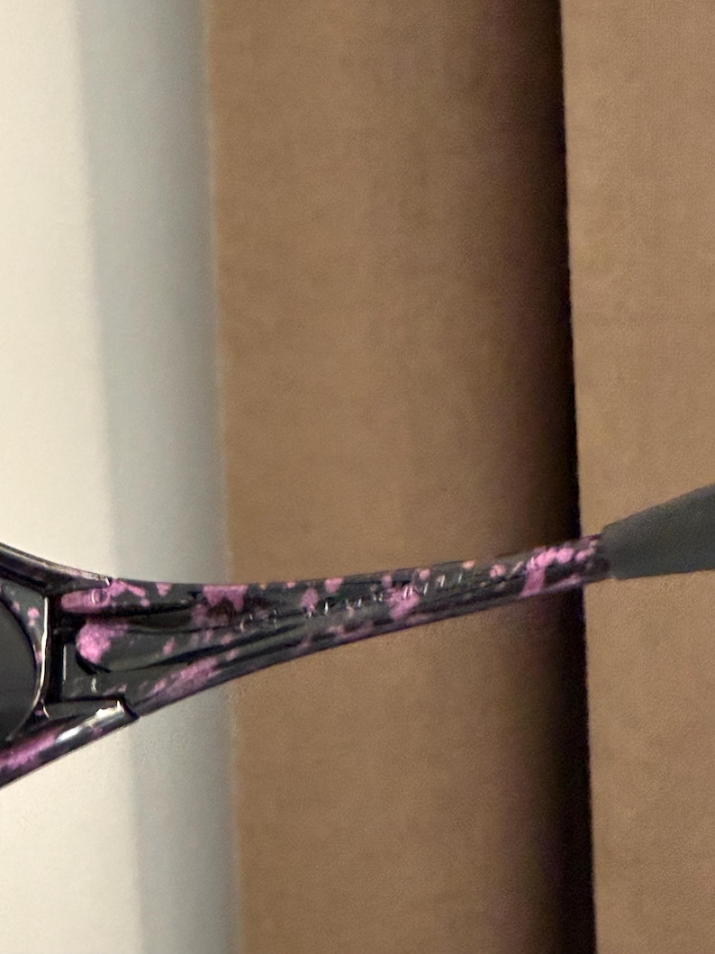 May include: Black sunglasses with a speckled pink and black pattern on the arms. The sunglasses have a dark lens and a sleek, modern design. The arms are slightly curved for a comfortable fit.