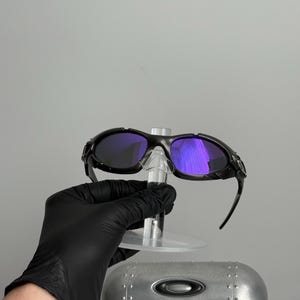 May include: Black sunglasses with purple lenses are displayed on a clear stand. The sunglasses have a sleek, sporty design. A silver case with the Oakley logo is visible below the sunglasses.
