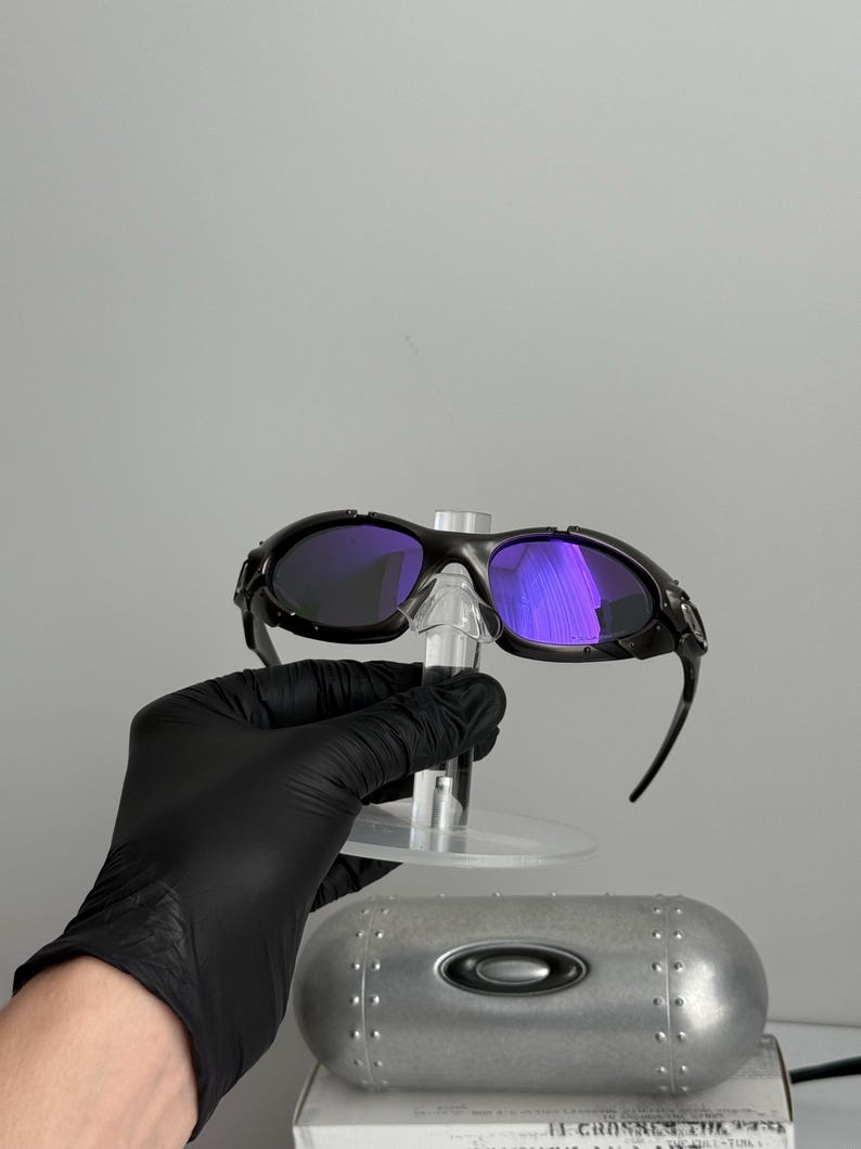 May include: Black sunglasses with purple lenses are displayed on a clear stand. The sunglasses have a sleek, sporty design. A silver case with the Oakley logo is visible below the sunglasses.