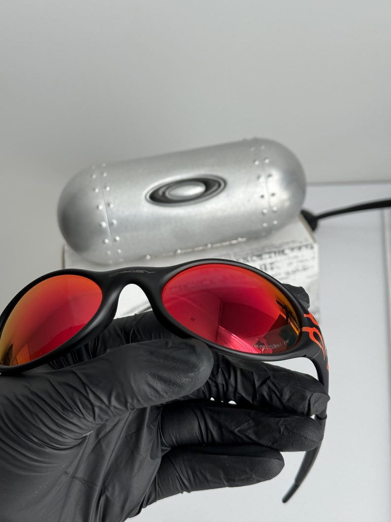 May include: Black sunglasses with red lenses and a flame design on the temples. The sunglasses are held in a black gloved hand. A silver case with the Oakley logo is in the background.
