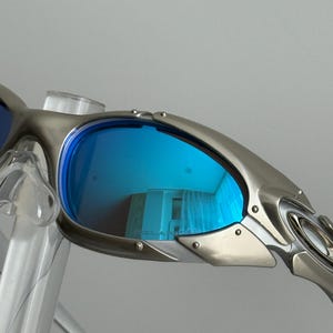 May include: Close-up of silver-framed sunglasses with blue lenses. The lenses reflect a room with curtains. The sunglasses have a sleek, modern design with visible rivets and a curved temple.