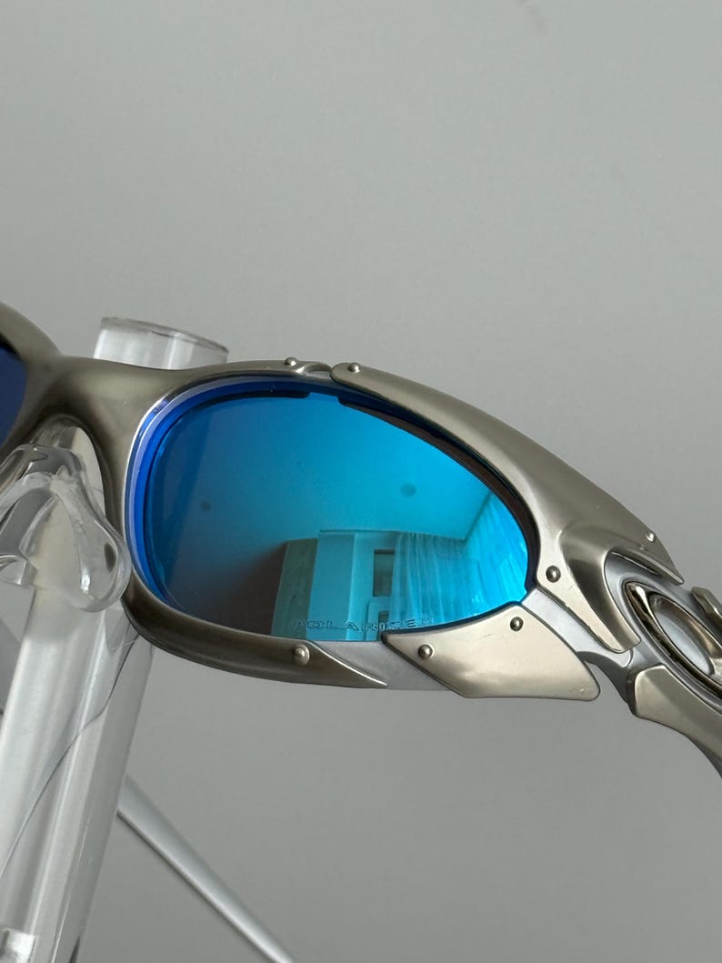 May include: Close-up of silver-framed sunglasses with blue lenses. The lenses reflect a room with curtains. The sunglasses have a sleek, modern design with visible rivets and a curved temple.