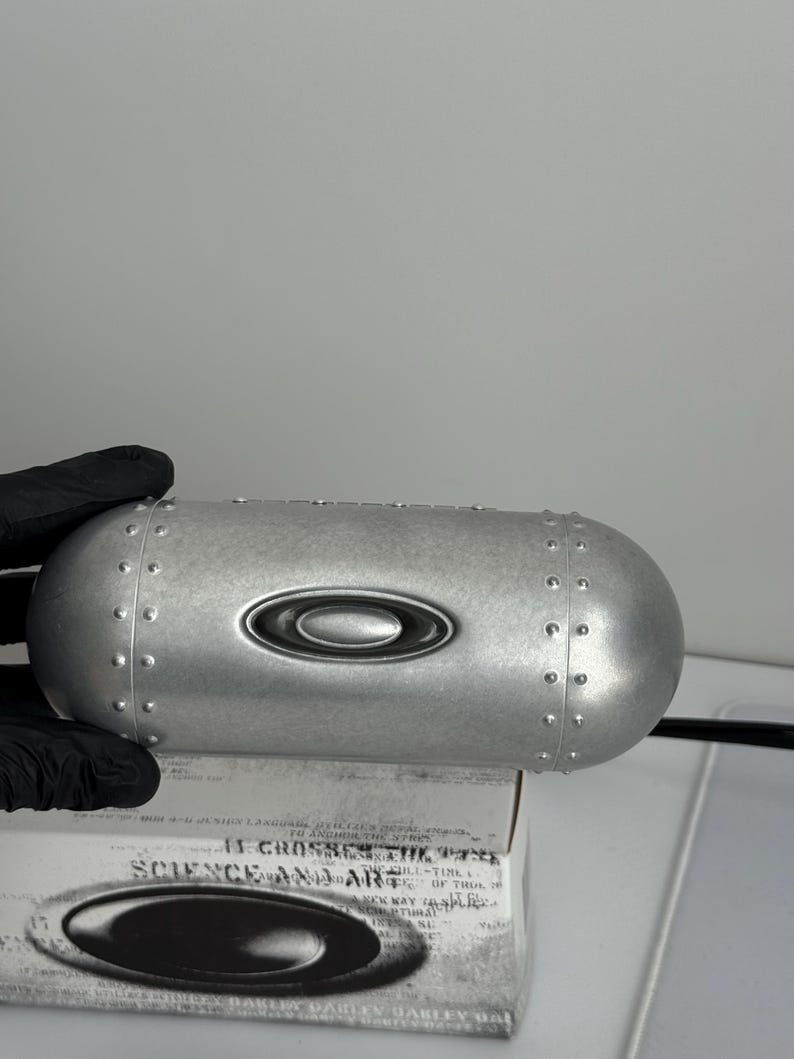 May include: A silver-colored, oval-shaped case with a riveted design and the Oakley logo. The case is resting on a box with the text "SCIENCE AND ART". The case is held by a gloved hand.