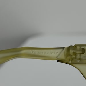 May include: Close-up of olive-green sunglasses. The text "PLANTARIS" is printed on the temple arm. The sunglasses have a modern, minimalist design.