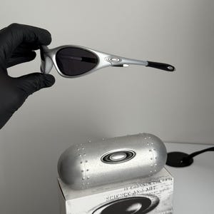 Puede incluir: Gafas de sol plateadas con lentes oscuras, sostenidas por una mano enguantada. Las gafas tienen un dise&ntilde;o elegante. Tambi&eacute;n se ven un estuche plateado y una caja con el texto "SCIENCE AND ART".
