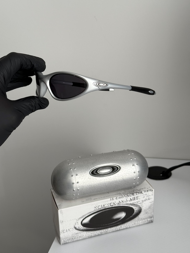 Puede incluir: Gafas de sol plateadas con lentes oscuras, sostenidas por una mano enguantada. Las gafas tienen un dise&ntilde;o elegante. Tambi&eacute;n se ven un estuche plateado y una caja con el texto "SCIENCE AND ART".