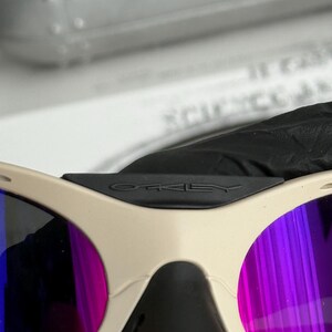 Może przedstawiać: Zbliżenie okular&oacute;w przeciwsłonecznych Oakley z beżową oprawką i fioletowo-niebieskimi lustrzanymi soczewkami. Czarny mostek ma logo Oakley. Okulary przeciwsłoneczne mają sportowy design.