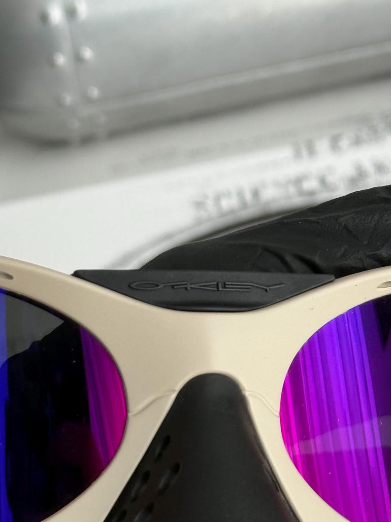 Może przedstawiać: Zbliżenie okular&oacute;w przeciwsłonecznych Oakley z beżową oprawką i fioletowo-niebieskimi lustrzanymi soczewkami. Czarny mostek ma logo Oakley. Okulary przeciwsłoneczne mają sportowy design.