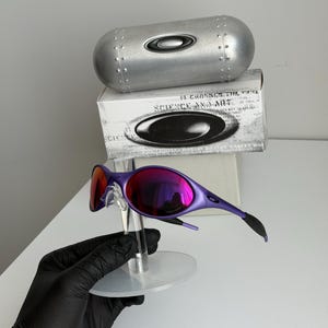 May include: Purple sunglasses with mirrored lenses, displayed on a clear stand. The glasses have a sleek, modern design. A silver case and a box with the text "SCIENCE AND ART" are in the background.