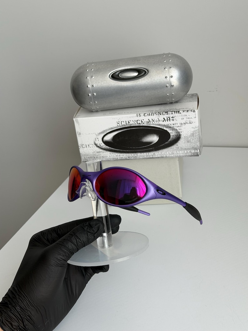 May include: Purple sunglasses with mirrored lenses, displayed on a clear stand. The glasses have a sleek, modern design. A silver case and a box with the text "SCIENCE AND ART" are in the background.