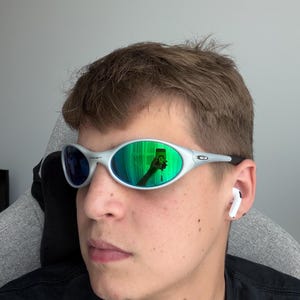 May include: Silver-framed sunglasses with green and blue reflective lenses. The sunglasses have a sleek, oval design. A person is wearing the sunglasses and a black shirt. White earbuds are in the person's ears.
