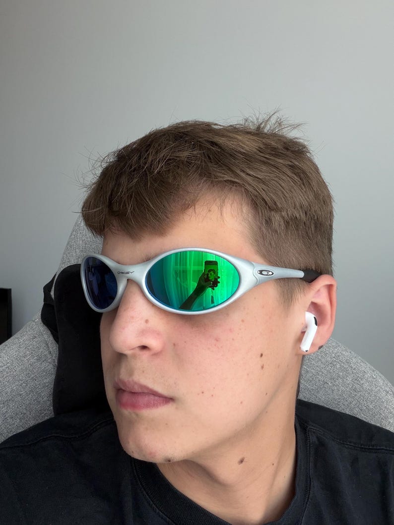 May include: Silver-framed sunglasses with green and blue reflective lenses. The sunglasses have a sleek, oval design. A person is wearing the sunglasses and a black shirt. White earbuds are in the person's ears.