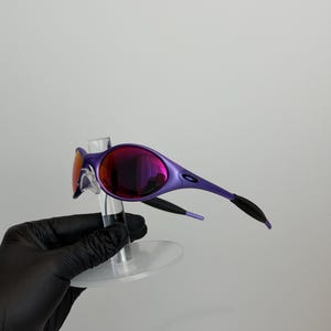 May include: Purple sunglasses with a sleek, modern design. The lenses have a mirrored finish, reflecting shades of red and purple. The frames are a vibrant purple, with black accents on the temples. The sunglasses are displayed on a clear stand.