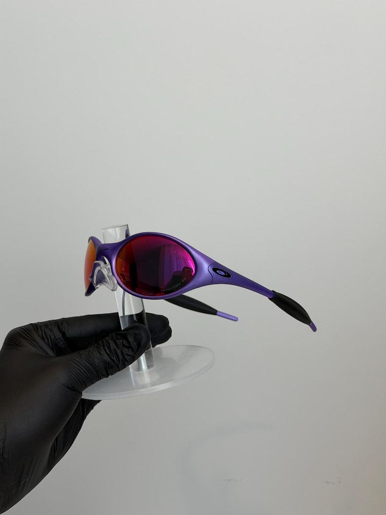 May include: Purple sunglasses with a sleek, modern design. The lenses have a mirrored finish, reflecting shades of red and purple. The frames are a vibrant purple, with black accents on the temples. The sunglasses are displayed on a clear stand.