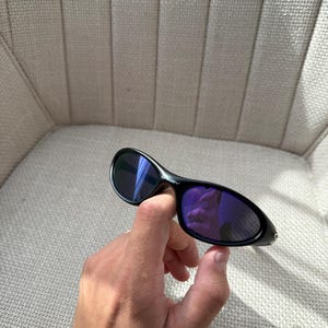 May include: Black sunglasses with purple lenses. The sunglasses have a curved design and are held in a hand. The lenses reflect the surroundings. The frame is black.
