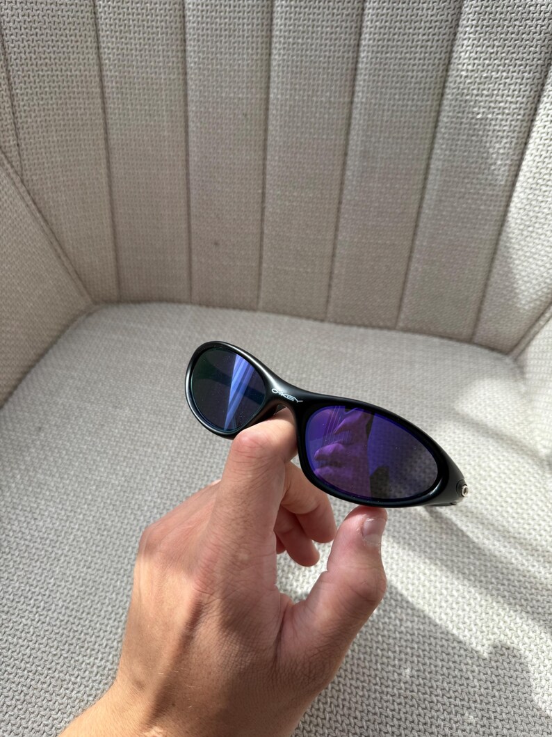 May include: Black sunglasses with purple lenses. The sunglasses have a curved design and are held in a hand. The lenses reflect the surroundings. The frame is black.