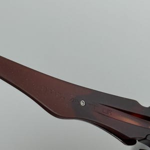 May include: Close-up of a brown and black patterned eyeglass arm. The arm has text and a small silver screw. The glasses are set against a light gray background.